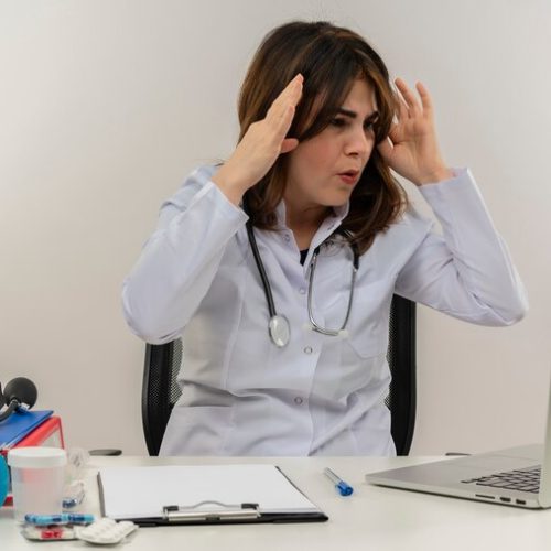 surprised-middle-aged-female-doctor-wearing-medical-robe-stethoscope-sitting-desk-with-medical-tools-clipboard-looking-laptop-touching-head-isolated_141793-69187